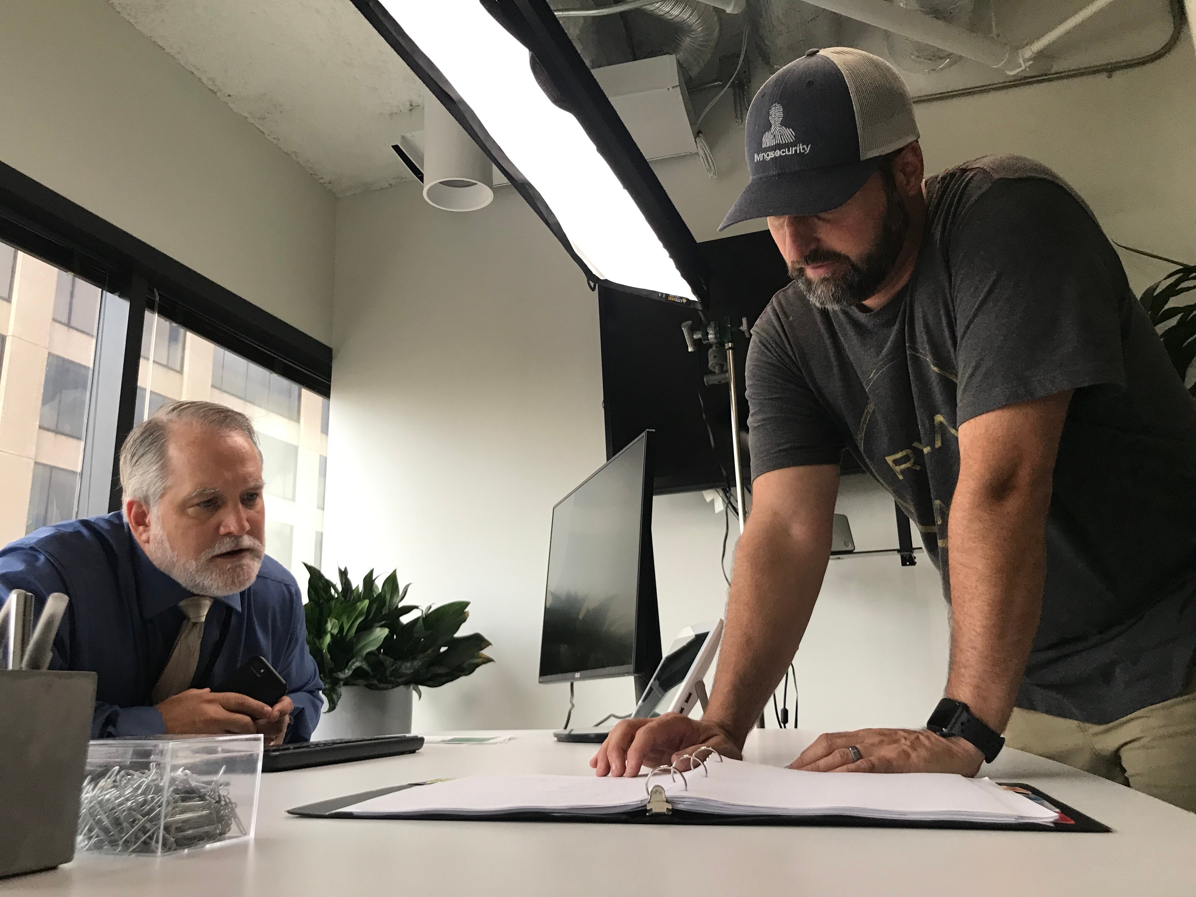 Image shows David West leaning over a table, looking at a binder, as an actor looks on. In the frame, we see elements of the set for True Eye, as well as lighting equipment.