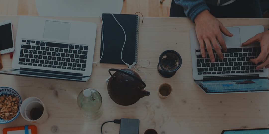 Desk with laptops, teapot and cup of tea, headphones, notebook and various electronic devices.