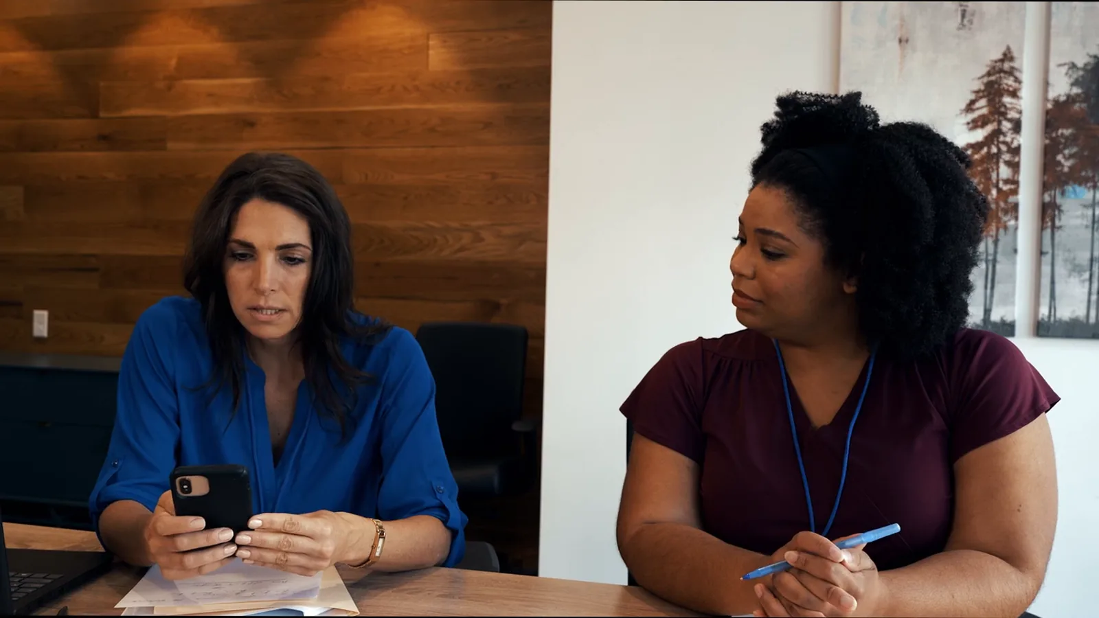Two employees of the fictional company sit at a table during the witness interviews, one checking her phone.